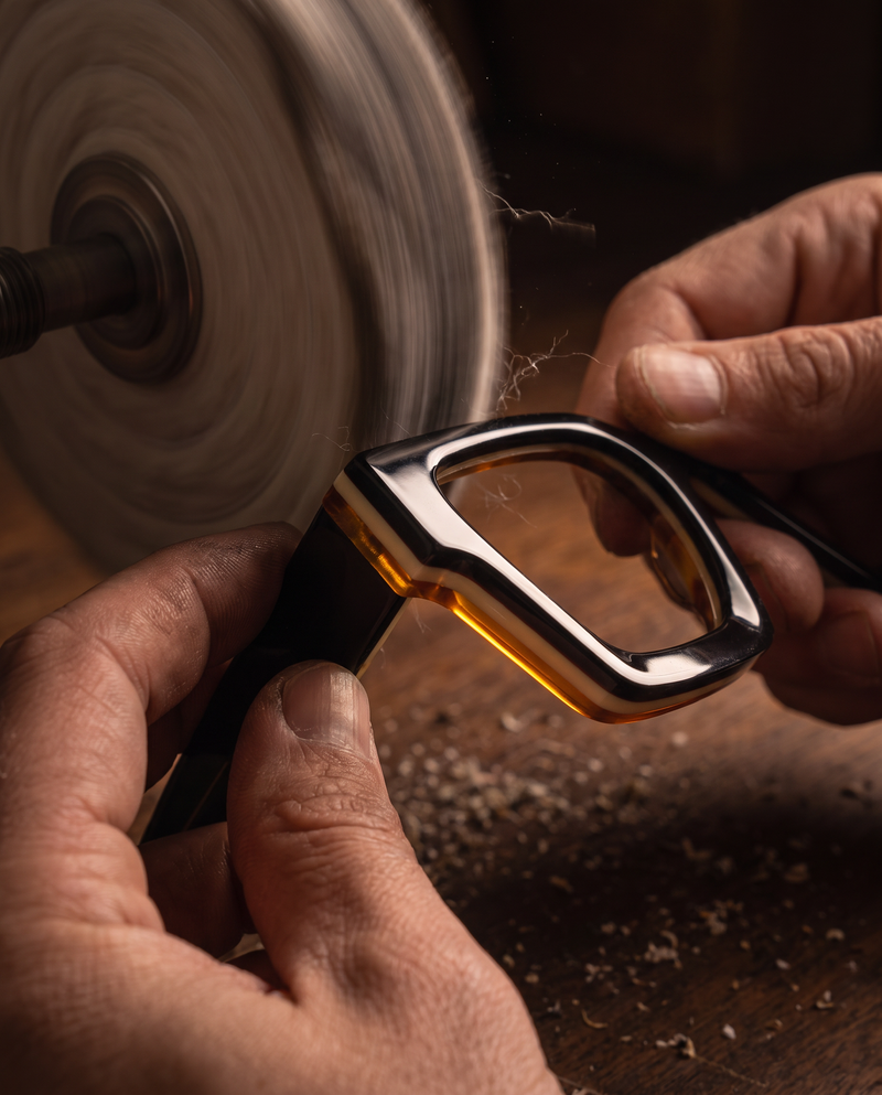 Hands polishing the layered edge of a square acetate sunglasses frame on a buffing wheel