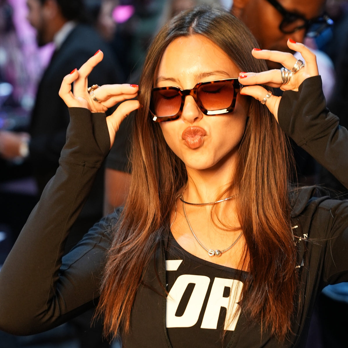 A woman posing in a crowded indoor event, wearing oversized square tortoiseshell sunglasses, a black zip-up hoodie with white graphic lettering, and a black belt bag.